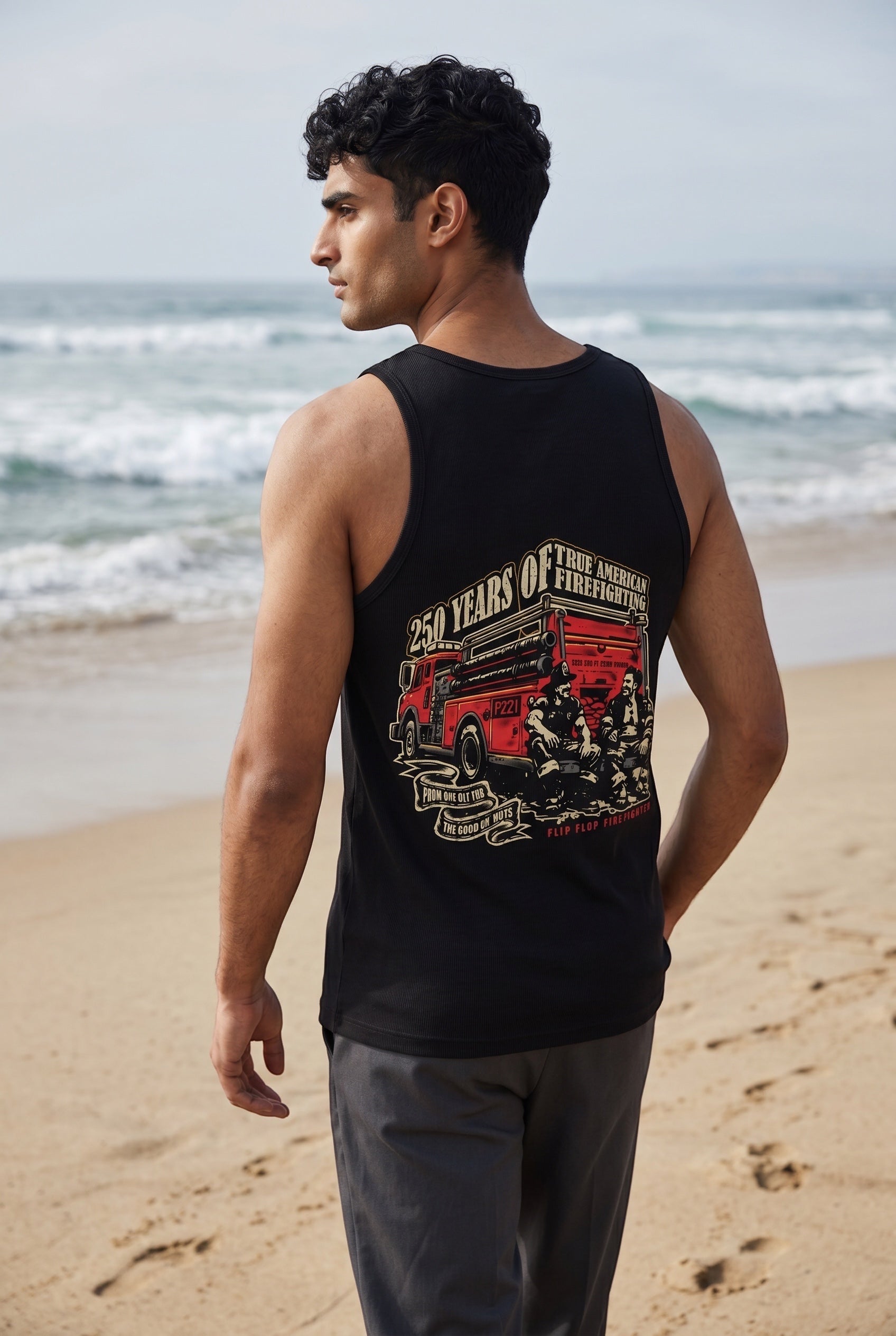 Flip Flop Firefighter black tank top with America 250 firefighting graphic on back, worn by a man on the beach.